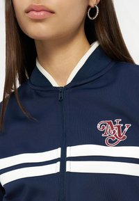 Νεαρή γυναίκα που φοράει μπλε σκούρο μπουφάν με φερμουάρ και άσπρες ρίγες, με κεντημένο λογότυπο "NY" στο στήθος και ασημένιο σκουλαρίκι κρίκο ορατό στο αριστερό αυτί.