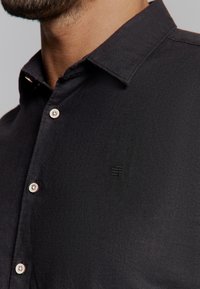 Zwarte button-up shirt van een gestructureerde stof, met een puntkraag en witte knopen. Klein logo geborduurd op de borst.