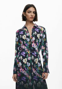 Schwarzes Hemdkleid mit Blumenmuster, ausgestattet mit langen Ärmeln, Kragen und Taillenbindeband. Die Farben umfassen Lila, Weiß, Pink und Grün. Fließender Stoff.