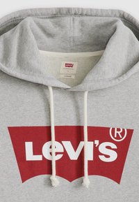 Felpa grigia in cotone con un grande logo rosso della Levi's. Presenta un cappuccio con lacci e una consistenza morbida. L'etichetta mostra la taglia "L" e i dettagli del marchio.