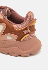 Smeđe i ružičaste tenisice s mrežastim panelima, teksturiranim gumenom potplatom, logotipom krokodila i žutim akcentima na peti. Sigurno prianjanje s petnim nastavkom.