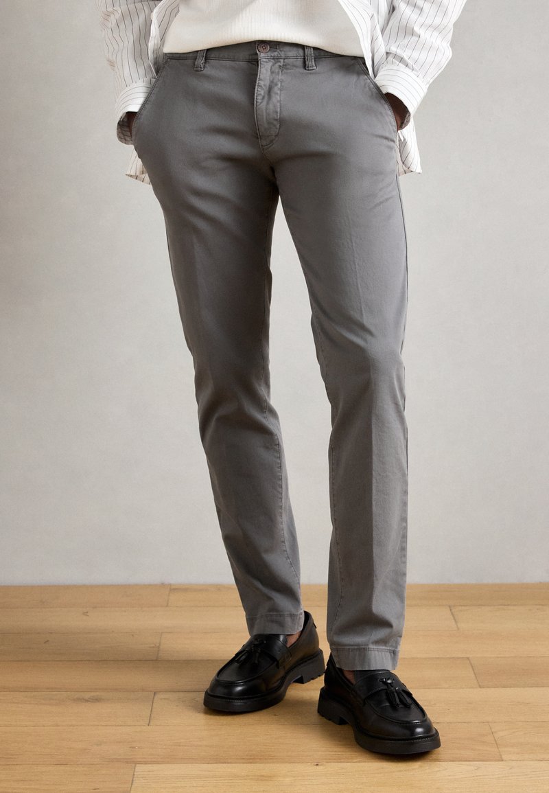 Pantalons en coton gris avec une coupe droite, dotés de poches latérales et d'une fermeture à bouton. Portés avec des mocassins noirs, exposés sur un sol en bois.