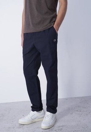 Mann trägt marineblaue Hose und weiße Sneaker, steht mit einer Hand in der Tasche vor schlichtem Hintergrund.