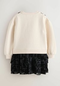 Cremfarbener Pullover mit langen Ärmeln und geripptem Saum, kombiniert mit einem mehrlagigen schwarzen Rüschenrock mit floralem Muster und Logo-Akzenten.