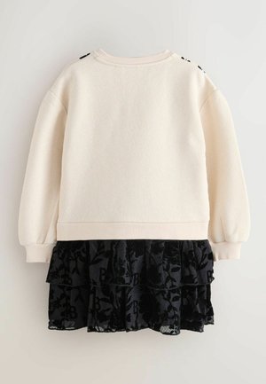 Krémový svetr s dlouhými rukávy a žebrovaným lemem, který je kombinován s vícerech černou sukní s volány, jež má květinový vzor a logo akcenty.