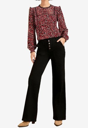 Mujer con blusa roja con estampado y mangas largas, pantalones negros de cintura alta y pierna ancha, y zapatos beige puntiagudos, mano en el bolsillo.