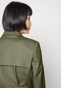 Kobieta z krótkimi czarnymi włosami, nosząca oliwkowy trencz, przedstawiona z boku i z tyłu na jednolitym tle.