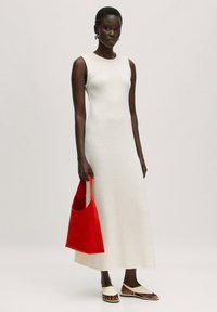 Witte mouwloze jurk met een gestructureerde afwerking, gecombineerd met een rode tote bag en beige platte sandalen, weergegeven tegen een neutrale achtergrond.