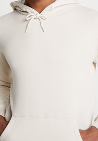 Beżowa bluza z kapturem wykonana z miękkiego materiału, wyposażona w kaptur ze sznurkiem, kieszeń kangurkę oraz ściągacze na rękawach. Gładki, minimalistyczny design.