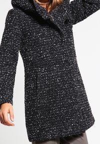 Cappotto a pois bianco e nero con cappuccio, maniche lunghe, tasche frontali e un tessuto testurizzato. Design classico con vestibilità su misura.