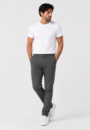 Mann mit dunklen Haaren, der ein schlichtes weißes T-Shirt, dunkelgraue Hose und weiße Turnschuhe trägt, steht mit den Händen in den Taschen.