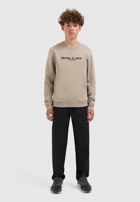Beige sweatshirt met een zwart "BALLIN"-logo, ronde hals, geribde manchetten, gecombineerd met zwarte broek en sportschoenen met een gestructureerde zool.