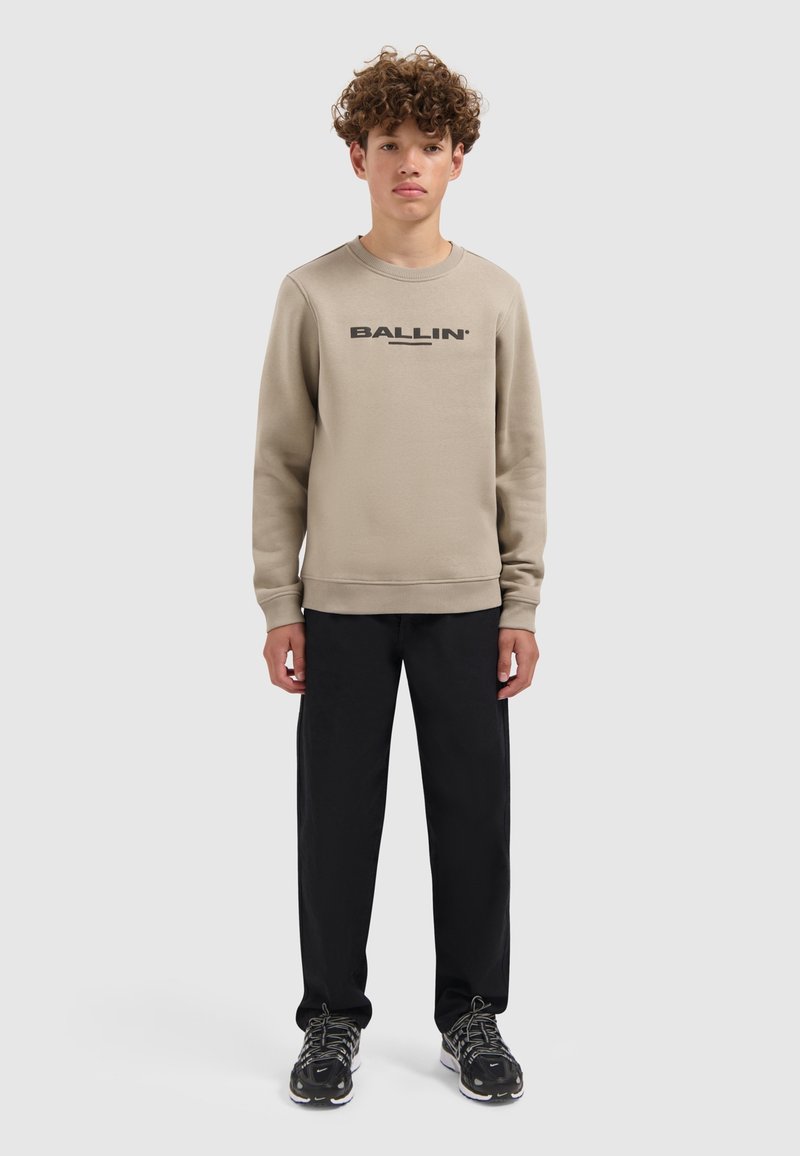Beige sweatshirt met een zwart "BALLIN"-logo, ronde hals, geribde manchetten, gecombineerd met zwarte broek en sportschoenen met een gestructureerde zool.