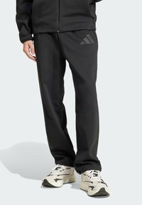 Čierne športové nohavice s hladkou textúrou, s tonálnym logom na ľavom stehne, skombinované so svetlými teniskami.