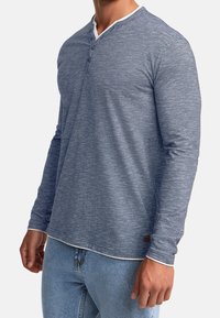 Long-sleeve, henley shirt in heathered blue fabric with white collar accent, featuring three buttons and a small logo tag at the hem.