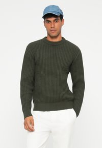 Maglione lavorato a maglia verde scuro con un motivo testurizzato a nido d'ape e polsini a coste. Abbinato a un cappellino blu chiaro e pantaloni bianchi.