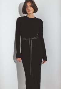 Črna rebrasta dolga obleka z oprijetim silhueto, poudarjena s kontrastnim pasom in podolgovatimi rokavi s režami.