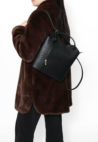 Borsa in pelle nera con texture perforata, tasca con zip e tracolla regolabile, indossata sopra un cappotto marrone peloso.
