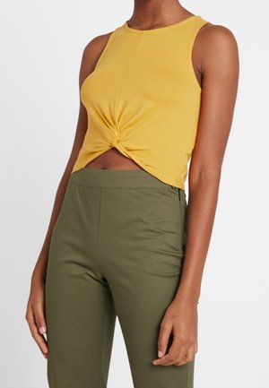 Gelbes ärmelloses Crop-Top mit einem geknoteten Frontdetail, kombiniert mit olivgrünen, hoch taillierten Hosen. Glatter Stoff, taillierte Passform.