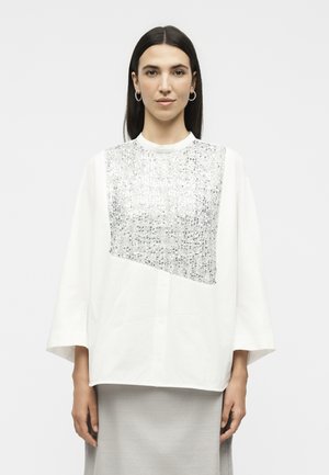 Kvinde med glat mørkt hår iført øreringe og en hvid oversized top med et stort sølvglitrende panel, stående foran en ensfarvet baggrund.