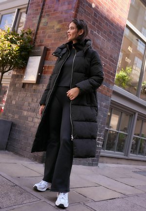 Doudoune noire avec une capuche et une fermeture éclair devant, associée à un pantalon noir ajusté et des baskets blanches. Décor urbain avec un mur en briques en arrière-plan.