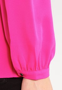 Blusa lunga a maniche con ruches, di colore rosa brillante, realizzata in tessuto morbido, con un accento di bottone nero alla fine della manica.