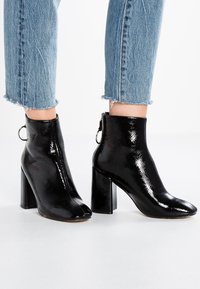 Schwarze Lackleder Stiefeletten mit glänzender Oberfläche, runder Spitze und klobigem Blockabsatz, ausgestattet mit einem Reißverschluss und einer Zuglasche.