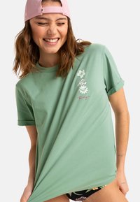 Mintgrøn bomuld t-shirt med korte ærmer og afslappet pasform. Designet omfatter blomstergrafik og skript detaljer i kontrasterende farver.