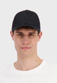 Gorra de béisbol negra con visera curva, con una superficie texturizada y un logo bordado en la parte frontal. Llevada con una camisa blanca.