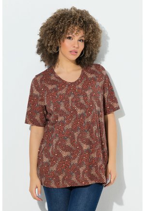 Frau mit lockigem Haar trägt ein braunes kurzärmeliges Oberteil mit Giraffenmuster und blaue Jeans, steht vor hellem Hintergrund.