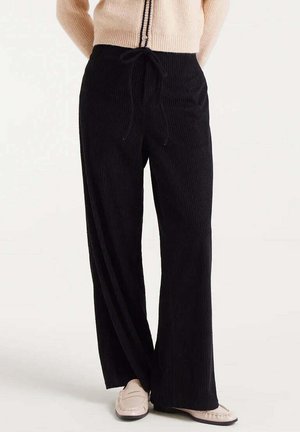 Mujer vestida con pantalones anchos negros con cintura elástica y mocasines beige, de pie contra un fondo blanco.