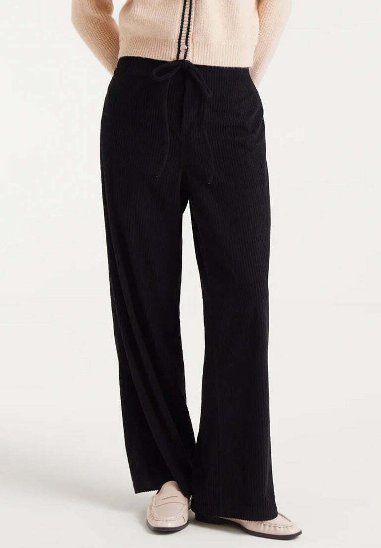 Mujer vestida con pantalones anchos negros con cintura elástica y mocasines beige, de pie contra un fondo blanco.