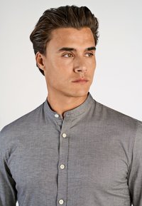 Giovane uomo con capelli scuri e acconciatura stilizzata, che indossa una camicia grigia con colletto a band, che guarda leggermente a destra su uno sfondo bianco.