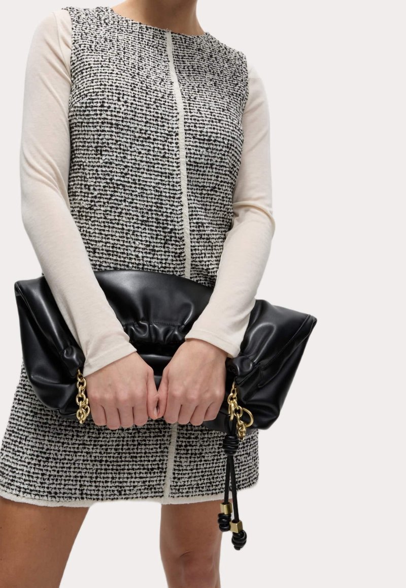 Femme portant une robe sans manches texturée noire et blanche par-dessus un haut crème à manches longues, tenant une grande pochette en cuir noir avec des détails en chaîne dorée.