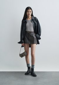 Camicia oversize in pelle nera, top grigio a costine, mini gonna marrone con cuciture a contrasto, stivali alla caviglia, calzini grigi, piccola borsa decorata.