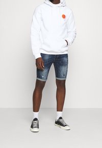 Valkoinen huppari, jossa on oranssi graafinen printti, yhdistettynä kuluneisiin farkkushortseihin, nilkkapituiset valkoiset sukat ja mustat tennarit.