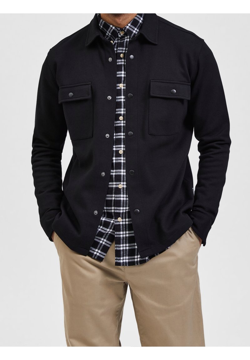 Homme portant un pantalon beige, une veste noire boutonnée avec poches à rabat, et une chemise à carreaux noir et blanc avec boutons.