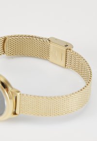 Montre en maille dorée avec un cadran rond. Le bracelet présente une texture de tissage fin et une boucle avec un texte gravé sur une surface polie.