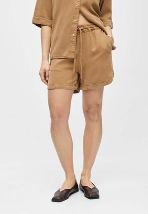 Mujer vestida con camisa marrón claro abotonada y pantalones cortos a juego con cordón, calzando zapatos planos marrón oscuro, de pie frente a un fondo liso.