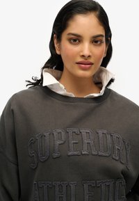 Sweat-shirt gris charbon avec "SUPERDRY ATHLETIC" brodé dans un fil plus foncé ; superposé sur une chemise blanche à col. Tissu doux et texturé.