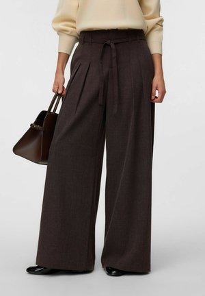 Persona que lleva pantalones anchos de tiro alto de color marrón oscuro con cinturón de tela, sosteniendo un bolso marrón oscuro y calzando zapatos negros.