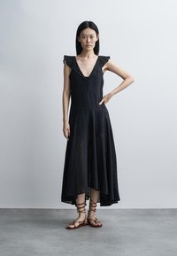 Žena s rovnými čiernymi vlasmi má na sebe čierne šaty bez rukávov s volánmi na ramenách a hnedé šnurovacie sandále na plochom chodidle, stojí na šedej podlahe.