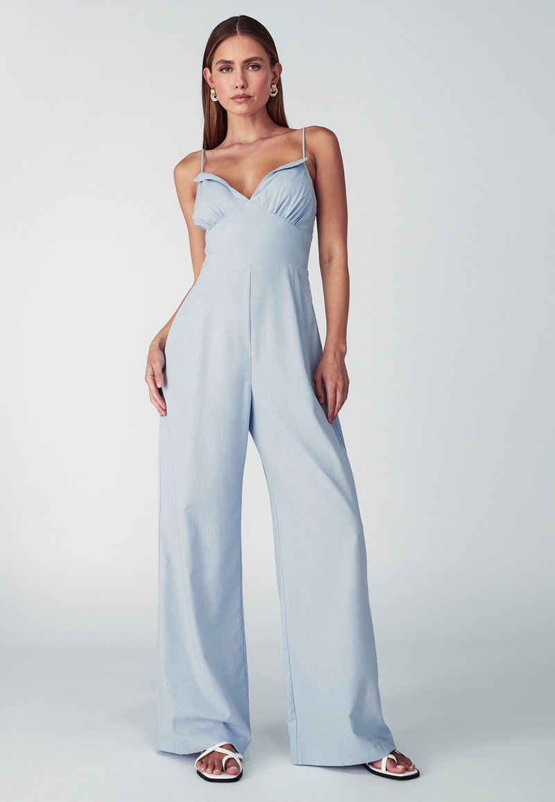 Mujer vestida con un mono sin mangas de pierna ancha en azul claro y sandalias blancas, de pie contra un fondo liso de color gris claro.