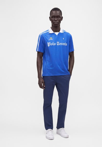 Homme debout portant un polo de tennis bleu, un pantalon bleu marine et des baskets blanches sur un fond blanc uni.