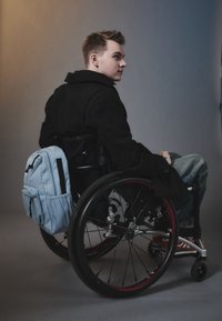 Black coat on a young man seated in a wheelchair, featuring red-accented wheels. He carries a light blue backpack on his side.