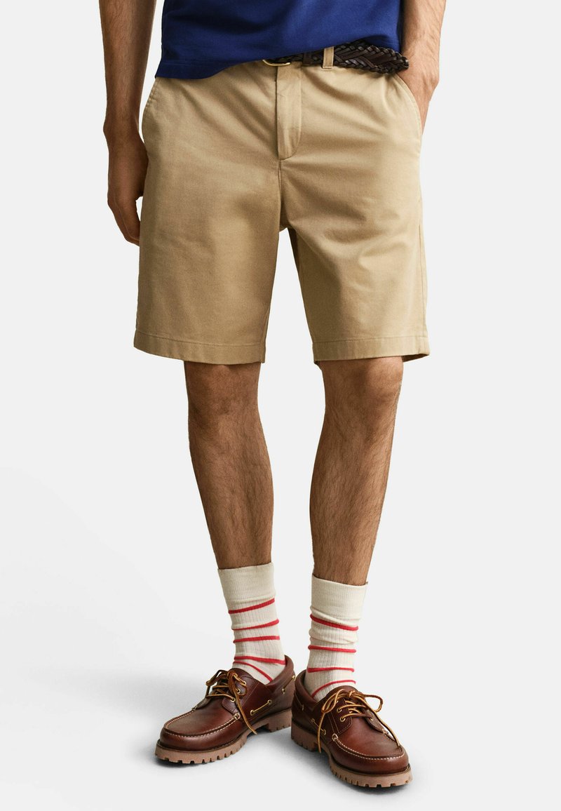 Homme portant un short beige, une ceinture tressée marron, des chaussettes blanches à rayures rouges et des chaussures en cuir marron, debout les mains dans les poches.