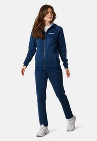 Navyblaues Sportjacken- und Hosen-Set aus strukturiertem Stoff, mit einem Reißverschluss vorne und verstellbarem Kapuzendesign, kombiniert mit weißen Sneakern.