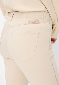 I jeans in denim beige presentano una texture liscia, tasche posteriori sottili e un dettaglio di cucitura argento a contrasto. L'etichetta del marchio riporta "RAPHAELA."