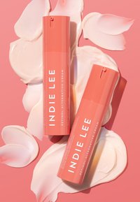 Kaksi korallinväristä INDIE LEE:n retinoli-vaihtoehtovoidepulloa vaaleanpunaisella taustalla, ympärillä kermamaisia värityksiä.