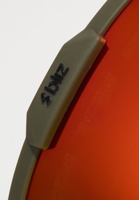 Bliz solbriller med en mørkeoliven ramme og store orange linser. Har en mat overflade og et fremtrædende sort logo på rammen.